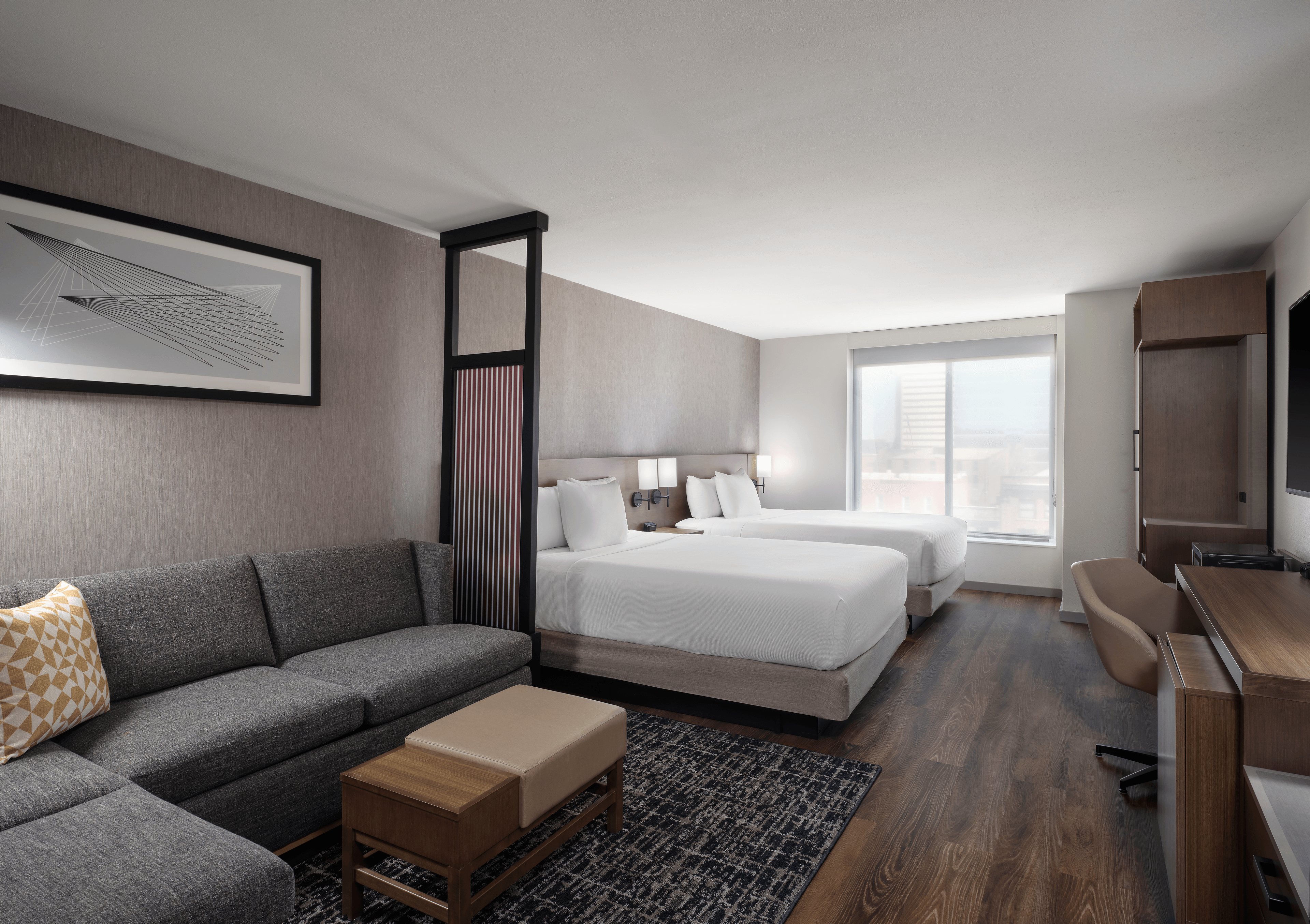 Modern hotel room with two queen beds, a grey sectional sofa, a wooden coffee table, and a work desk, featuring a large window with a cityscape view.